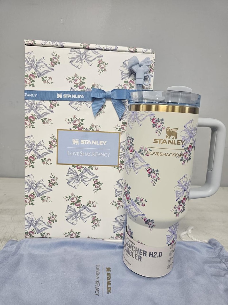 May include: A white Stanley Quencher H2.0 tumbler with a floral and bow design, matching a gift box. The tumbler has a handle and a lid. The box and tumbler feature the text "Stanley" and "LoveShackFancy".