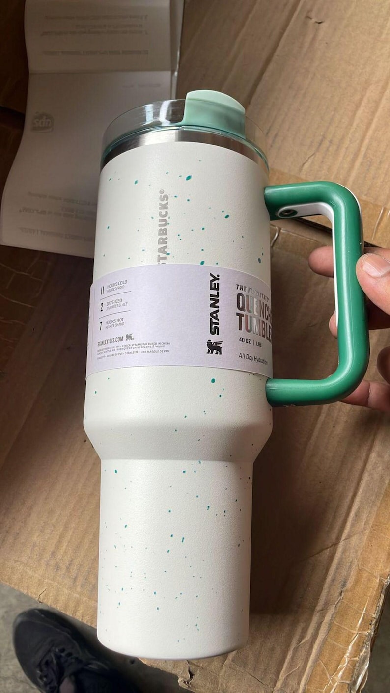 May include: A white ceramic mug with a black handle and the text "But First, Coffee" in a bold, black font. The mug is set against a blurred background, suggesting a cosy setting.