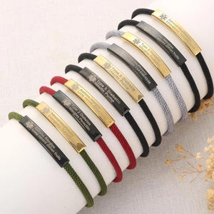Può includere: Una collezione di braccialetti di allerta medica con cordini di vari colori, tra cui nero, rosso e verde. Ogni braccialetto presenta una placca metallica rettangolare con informazioni mediche e simboli incisi. I braccialetti sono esposti su una superficie bianca.