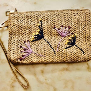 Può includere: Pochette rettangolare in paglia intrecciata con cerniera dorata e cinturino da polso. La pochette è decorata con ricami floreali viola, gialli e blu navy. La borsa ha una palette di colori neutri.