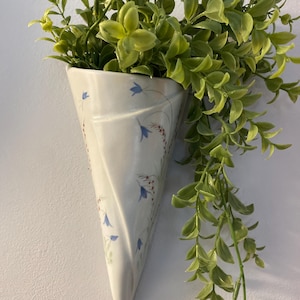 May include: A white, cone-shaped wall planter with a floral design of blue flowers and red berries. The planter is filled with green, leafy faux plants that cascade down the side. The planter is mounted on a white wall.