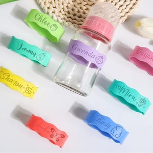 May include: A clear baby bottle with a pink lid and several colorful silicone bands. The bands are in various colors, each with a name and a small graphic, including "Chloe", "Jeremy", "Sharon", "Gwenda", "Dayana", "Mira", "Amelia", and "Kirsty".