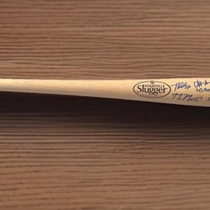 May include: A wooden Louisville Slugger baseball bat with signatures. The bat is light brown with the Louisville Slugger logo and text. The signatures are in black ink. The bat is lying on a brown wooden surface.