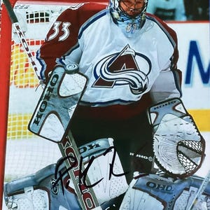 Może przedstawiać: Podpisane zdjęcie bramkarza hokejowego w stroju Colorado Avalanche. Bramkarz ma na sobie kask z kwiatowym wzorem, białą koszulkę z logo drużyny i bordowe akcenty. Widoczne są ochraniacze i kij.