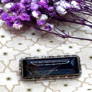 Könnte beinhalten: Eine rechteckige silberne Brosche mit einem dunkelblauen, fast schwarzen Stein in der Mitte. Die Brosche hat einen dekorativen Rand und ist vor einem Hintergrund aus lila und weißen Blumen platziert. Die Blumen sind auf einem gemusterten Stoff angeordnet.