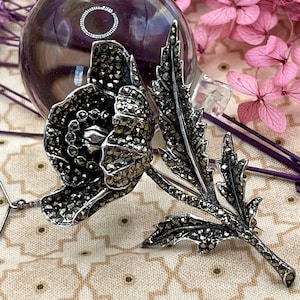 Op de afbeelding: Zilverkleurige markasiet broche in de vorm van een klaproos en bladeren. De broche heeft een gedetailleerd ontwerp met kleine, sprankelende steentjes. De klaproos heeft een veiligheidsspeldsluiting. De achtergrond omvat roze bloemen en een patroon.