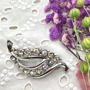 Può includere: Spilla argentata a forma di foglia con strass trasparenti e scintillanti. La spilla è posta su uno sfondo di tessuto bianco traforato, con accenti floreali viola e verdi.