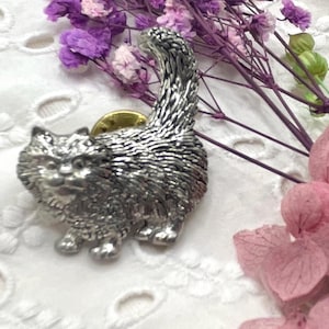Puede incluir: Broche de gato plateado con diseño texturizado, cola levantada y cierre dorado. El broche se encuentra sobre un fondo de flores moradas y rosas y tela de encaje blanco.