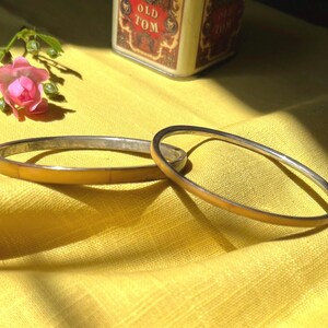 May include: Two circular bangles, one featuring a smooth yellow inlay and the other a thin metallic band with a yellow finish. These jewellery pieces are positioned together, showcasing their simple, rounded profiles and reflective surfaces.