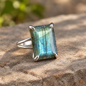 May include: A silver ring featuring a large, rectangular labradorite gemstone with a polished surface. The stone displays iridescent blue and green flashes within its structure. The metal band is split into two thin, polished silver prongs.