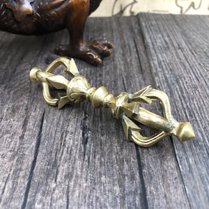 May include: An antique brass vajra, a ritual object, is displayed on a wooden surface. The vajra has detailed designs and a symmetrical shape, with pointed ends and a central decorative element. The background includes a bronze sculpture.