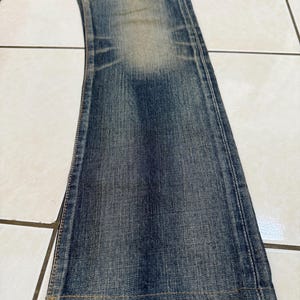 May include: Close-up of a pair of blue denim jeans. The jeans have a faded wash and a slight flare at the bottom. The fabric shows signs of wear and tear, with visible seams and stitching.