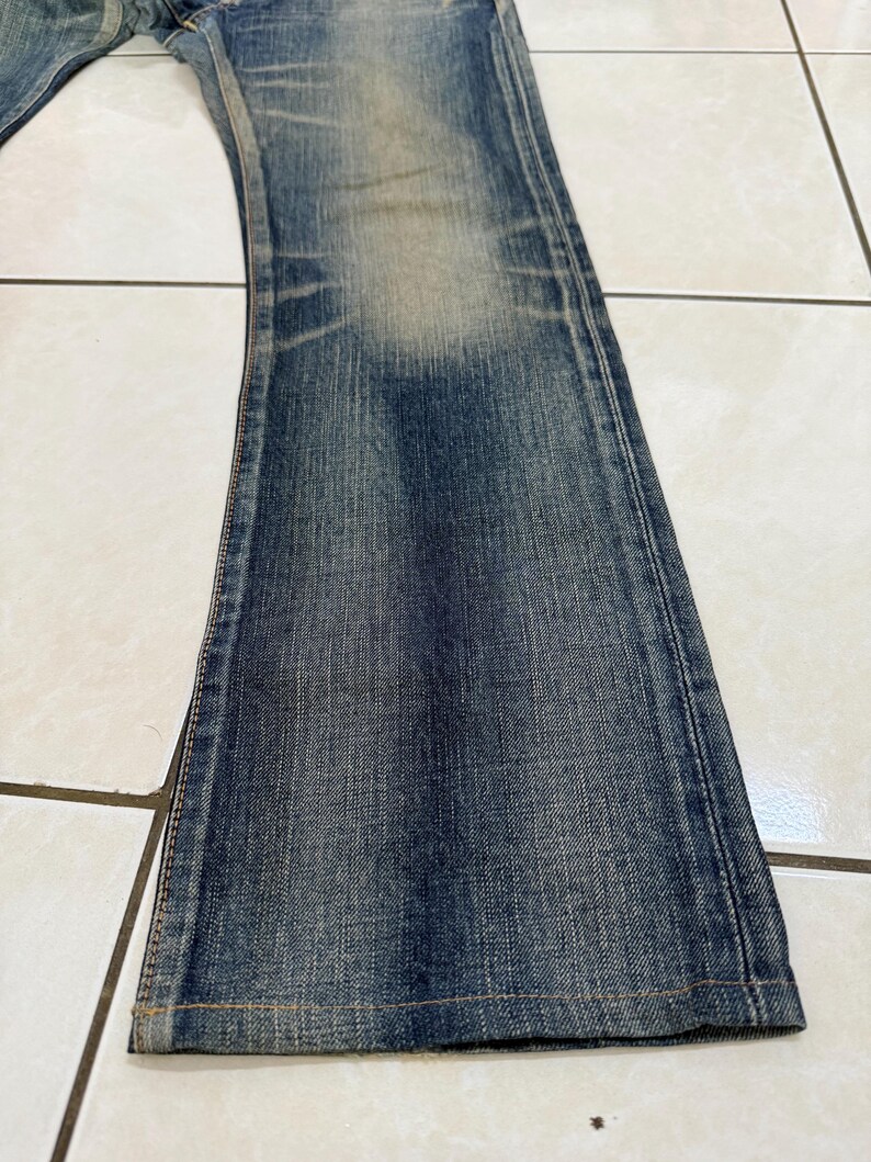 May include: Close-up of a pair of blue denim jeans. The jeans have a faded wash and a slight flare at the bottom. The fabric shows signs of wear and tear, with visible seams and stitching.