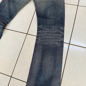 May include: Faded blue denim jeans with a worn appearance. The jeans have a classic five-pocket design, with visible wear and tear, including distressed areas and fading. The denim has a medium wash with darker shading on the legs.
