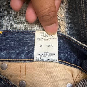 May include: Close-up of a denim garment's interior, showcasing a tag with Japanese text and care symbols. The tag indicates the fabric is 100% cotton. Silver rivets and beige fabric are also visible.