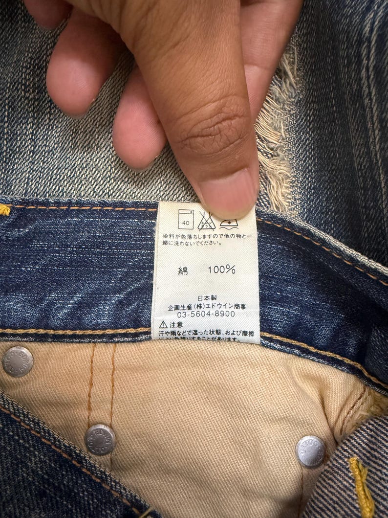 May include: Close-up of a denim garment's interior, showcasing a tag with Japanese text and care symbols. The tag indicates the fabric is 100% cotton. Silver rivets and beige fabric are also visible.