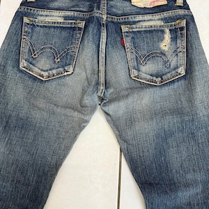 May include: Faded blue denim jeans with distressed details, featuring classic back pockets with signature stitching. A red Levi's tag is visible on one pocket. The jeans have a worn look, with a leather patch at the waistband.