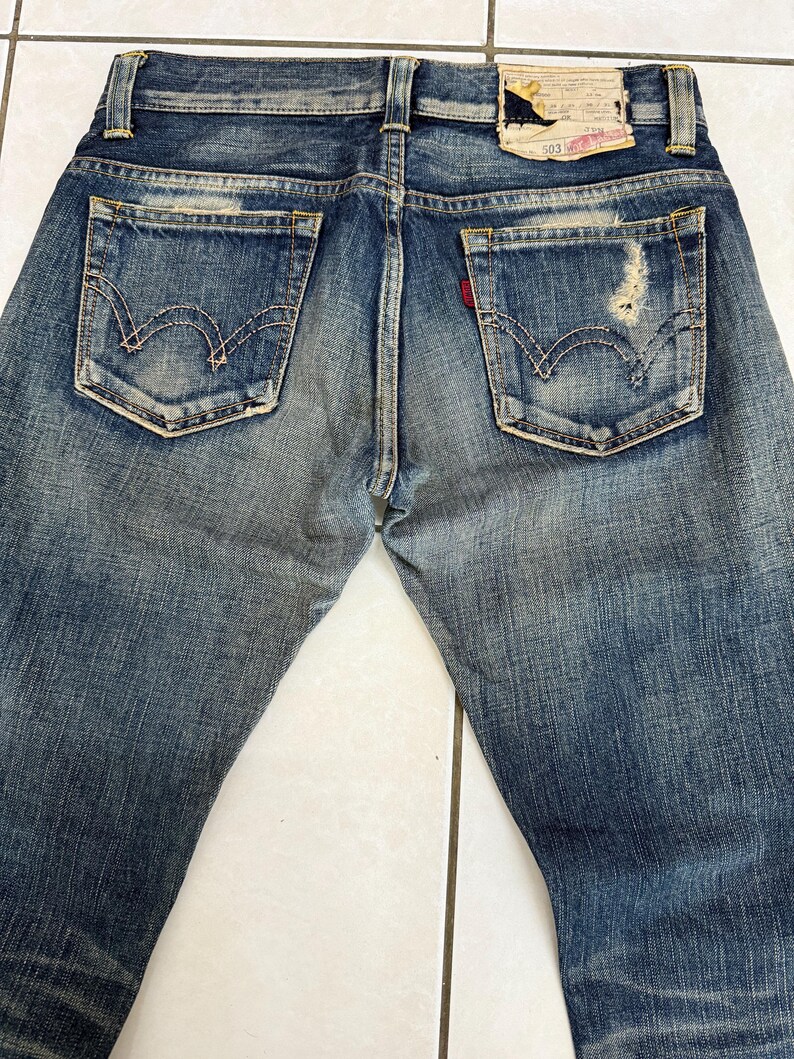 May include: Faded blue denim jeans with distressed details, featuring classic back pockets with signature stitching. A red Levi's tag is visible on one pocket. The jeans have a worn look, with a leather patch at the waistband.