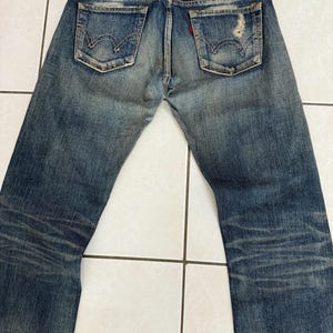 May include: Faded blue denim jeans with distressed details, featuring classic back pockets and a red tag. The jeans have a worn appearance, with visible wear marks and a vintage aesthetic.