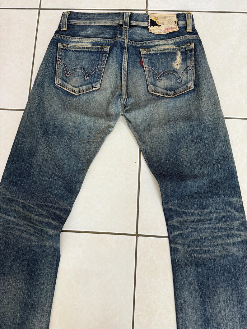 May include: Faded blue denim jeans with distressed details, featuring classic back pockets and a red tag. The jeans have a worn appearance, with visible wear marks and a vintage aesthetic.