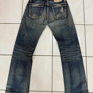 May include: Faded blue denim jeans with a distressed look. The jeans have two back pockets, a red tag, and a worn leather patch. The jeans are laid flat on a tiled surface.