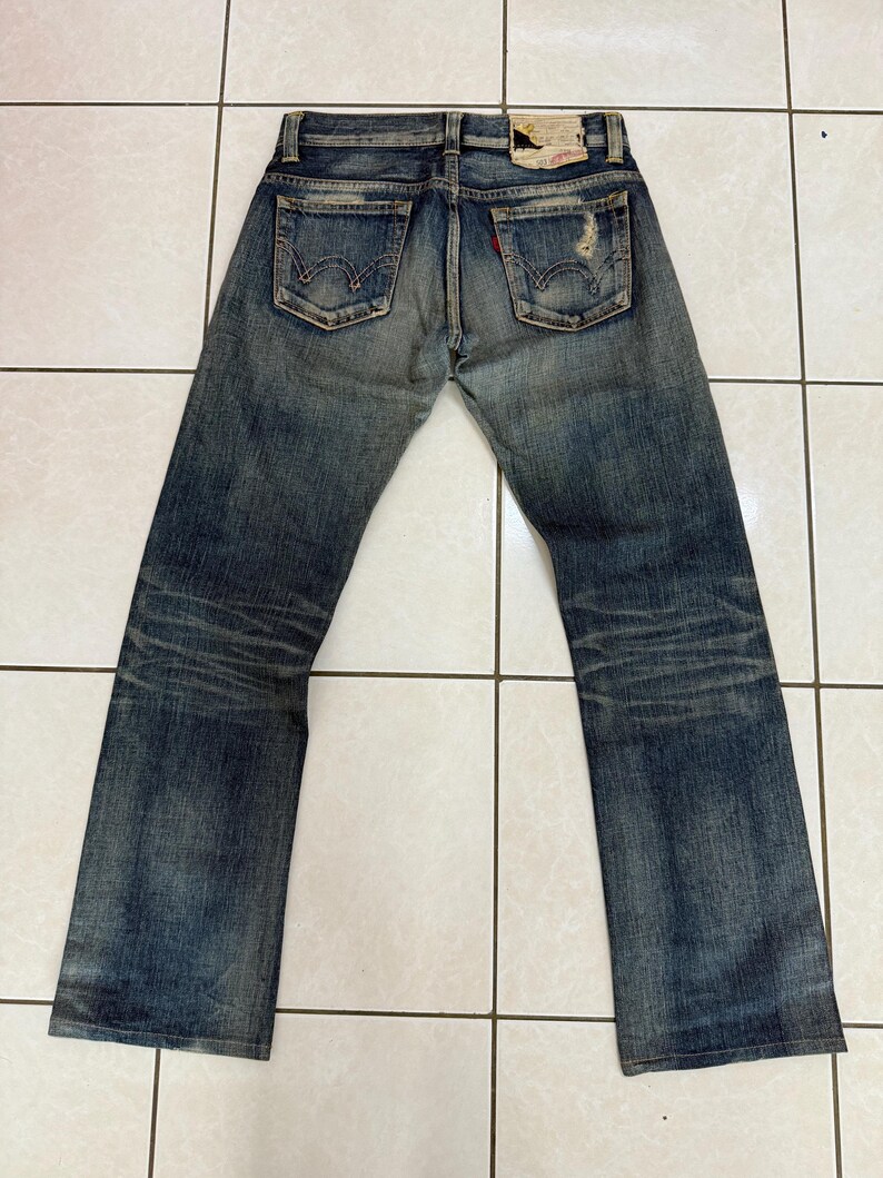 May include: Faded blue denim jeans with a distressed look. The jeans have two back pockets, a red tag, and a worn leather patch. The jeans are laid flat on a tiled surface.