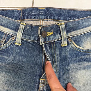 May include: Close-up of blue denim jeans with a button closure and zipper. The jeans have yellow stitching and metal rivets. The denim has a faded, worn appearance. The jeans are a classic style.