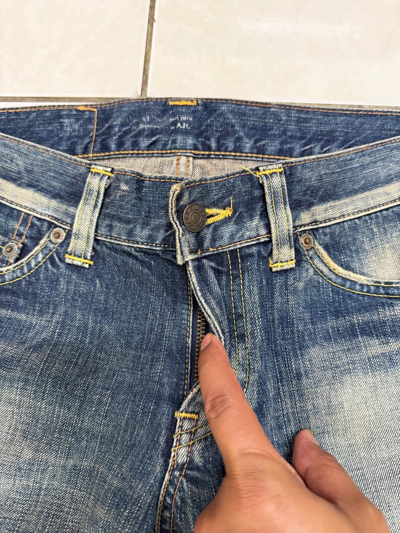 May include: Close-up of blue denim jeans with a button closure and zipper. The jeans have yellow stitching and metal rivets. The denim has a faded, worn appearance. The jeans are a classic style.