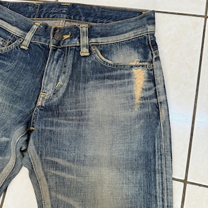 May include: Close-up of blue denim jeans with a distressed, worn appearance. The jeans feature a button closure, belt loops, and a classic five-pocket design. Fading and a tear on the front add to the vintage aesthetic. Yellow stitching is visible.