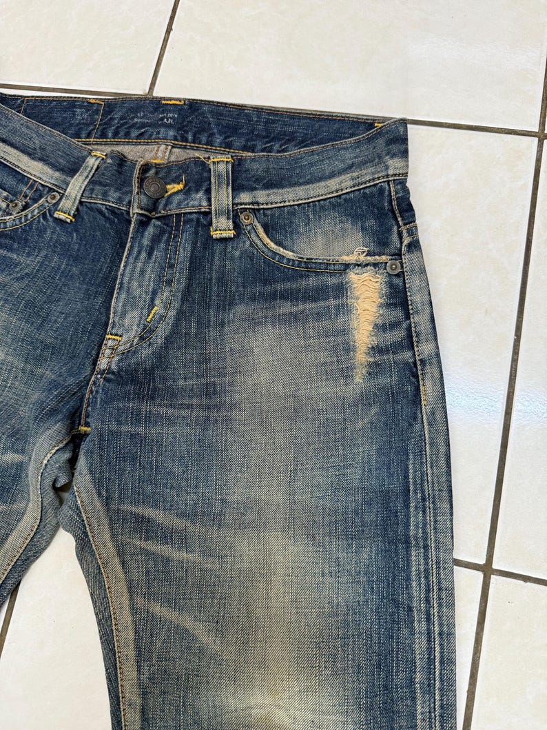 May include: Close-up of blue denim jeans with a distressed, worn appearance. The jeans feature a button closure, belt loops, and a classic five-pocket design. Fading and a tear on the front add to the vintage aesthetic. Yellow stitching is visible.