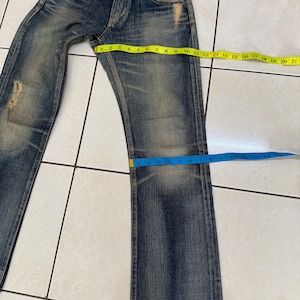 May include: Blue denim jeans with a distressed, worn look. The jeans have a button fly and multiple pockets. A yellow measuring tape shows a waist measurement of approximately 21 inches. The leg length is approximately 13 inches.