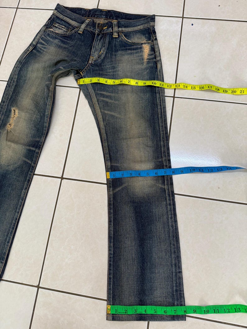 May include: Blue denim jeans with a distressed, worn look. The jeans have a button fly and multiple pockets. A yellow measuring tape shows a waist measurement of approximately 21 inches. The leg length is approximately 13 inches.