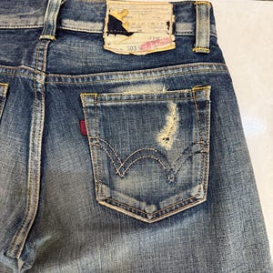May include: Close-up of the back of blue denim jeans, showing the back pocket with a distressed area. The jeans have a faded wash and a leather patch at the waistband. The pocket features the classic stitching design.