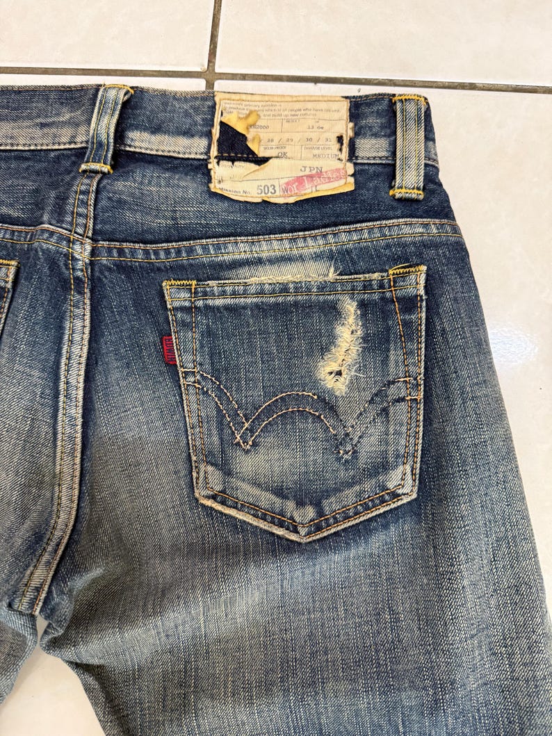 May include: Close-up of the back of blue denim jeans, showing the back pocket with a distressed area. The jeans have a faded wash and a leather patch at the waistband. The pocket features the classic stitching design.