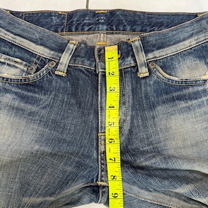 May include: Close-up of blue denim jeans with a yellow measuring tape showing a length of approximately 10 inches. The jeans have a button closure and classic five-pocket design. The denim has a faded, worn look.