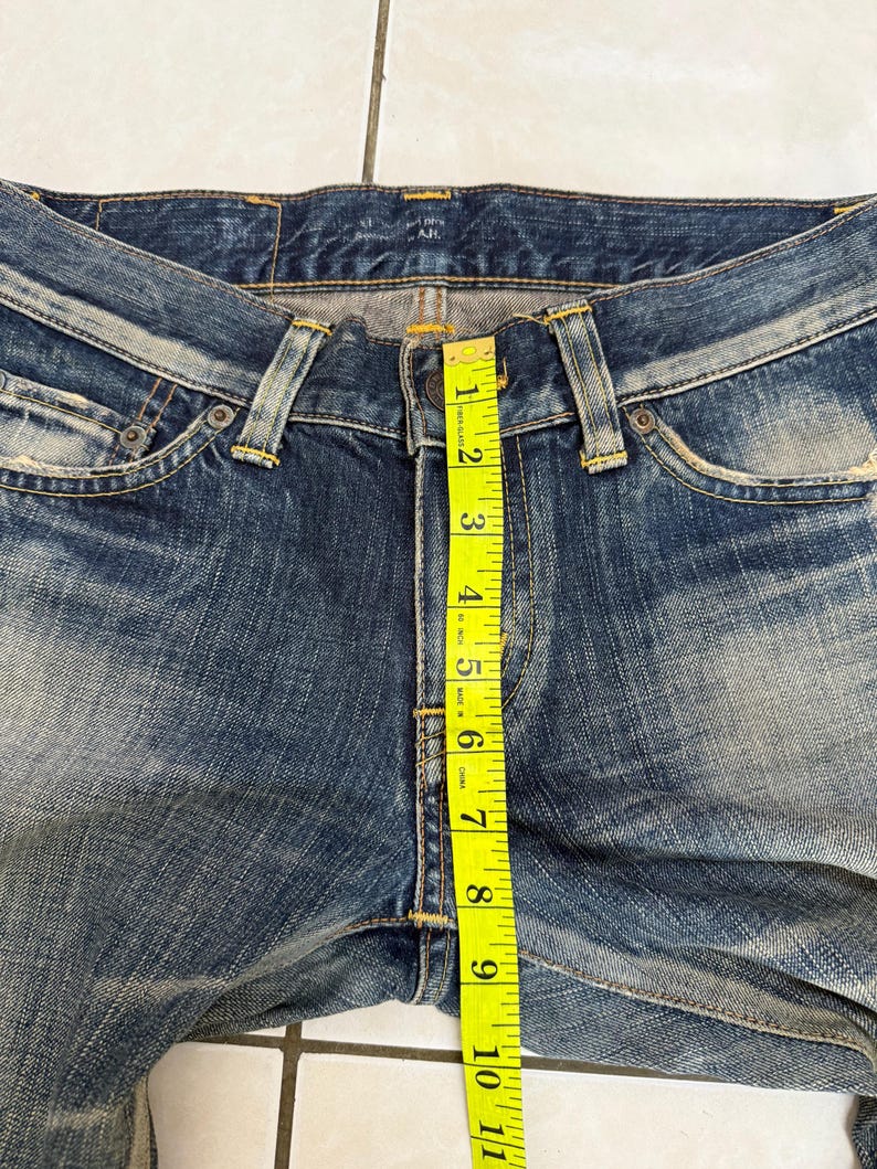 May include: Close-up of blue denim jeans with a yellow measuring tape showing a length of approximately 10 inches. The jeans have a button closure and classic five-pocket design. The denim has a faded, worn look.