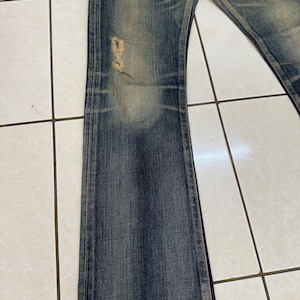 May include: Faded blue denim jeans with a distressed look. The jeans have a slight flare at the bottom and a ripped section on the left leg. The denim has a worn appearance with lighter patches.