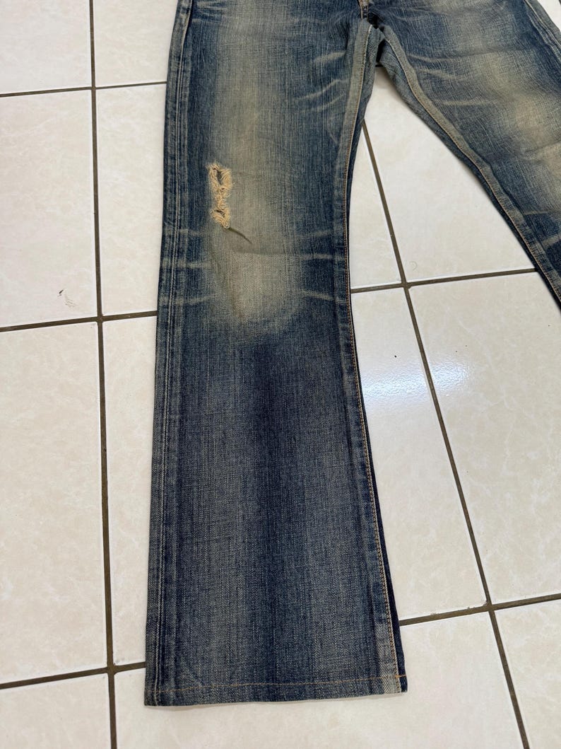 May include: Faded blue denim jeans with a distressed look. The jeans have a slight flare at the bottom and a ripped section on the left leg. The denim has a worn appearance with lighter patches.
