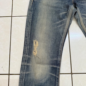 May include: Close-up of blue denim jeans with a distressed, worn appearance. The jeans have a faded wash and a ripped section on the left leg, revealing the underlying threads. The fabric shows signs of wear and tear, with visible fading and subtle variations in color.