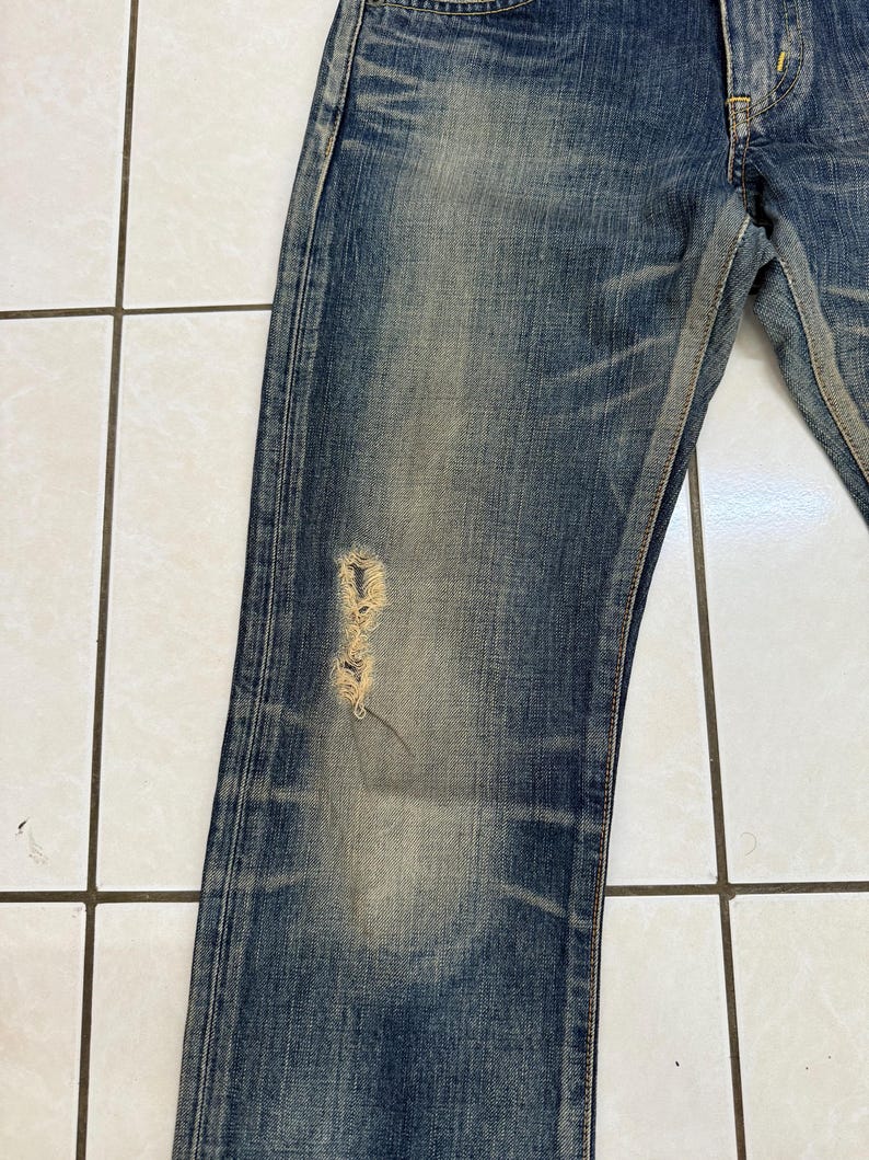 May include: Close-up of blue denim jeans with a distressed, worn appearance. The jeans have a faded wash and a ripped section on the left leg, revealing the underlying threads. The fabric shows signs of wear and tear, with visible fading and subtle variations in color.