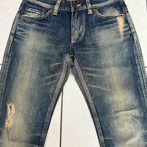 May include: Blue denim jeans with a distressed, faded appearance. The jeans feature a classic five-pocket design, a button and zipper closure, and visible wear and tear, including a ripped section on the left leg. The denim has a worn, vintage look.
