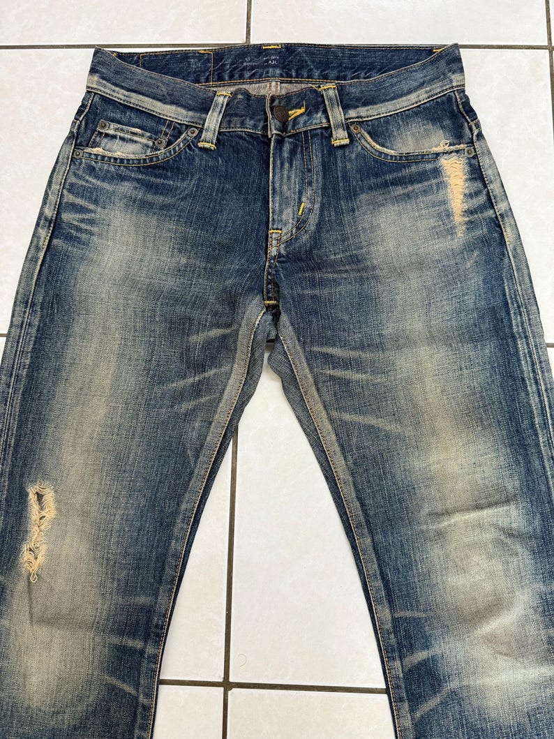 May include: Blue denim jeans with a distressed, faded appearance. The jeans feature a classic five-pocket design, a button and zipper closure, and visible wear and tear, including a ripped section on the left leg. The denim has a worn, vintage look.