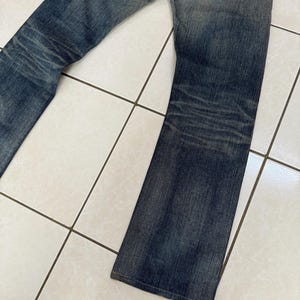May include: Faded blue denim jeans with a distressed look. The jeans have a classic five-pocket design, a button and zipper closure, and a slight flare at the bottom of the legs. The denim has a worn appearance with fading and whiskering.