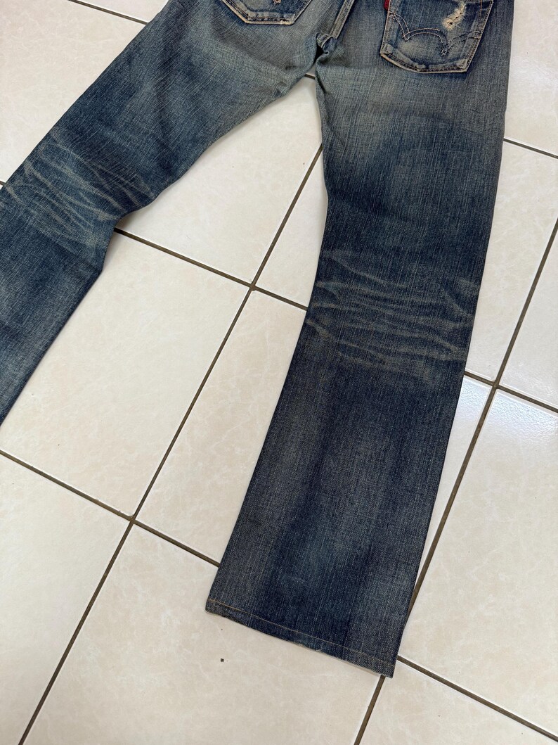 May include: Faded blue denim jeans with a distressed look. The jeans have a classic five-pocket design, a button and zipper closure, and a slight flare at the bottom of the legs. The denim has a worn appearance with fading and whiskering.