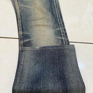 May include: Faded blue denim jeans with a folded cuff. The jeans show wear and tear with lighter patches and subtle distressing. The fabric has a textured appearance, and the overall look is casual and vintage.