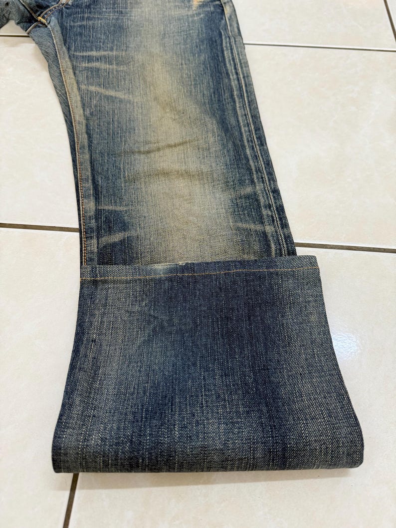 May include: Faded blue denim jeans with a folded cuff. The jeans show wear and tear with lighter patches and subtle distressing. The fabric has a textured appearance, and the overall look is casual and vintage.
