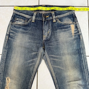 May include: Blue denim jeans with a distressed, faded appearance. The jeans have a classic five-pocket design, a button and zipper closure, and a yellow measuring tape across the waist. The waist measures approximately 18 inches.