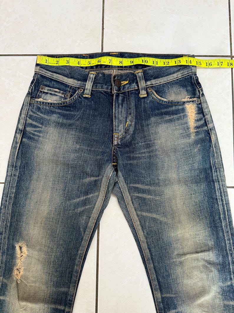 May include: Blue denim jeans with a distressed, faded appearance. The jeans have a classic five-pocket design, a button and zipper closure, and a yellow measuring tape across the waist. The waist measures approximately 18 inches.