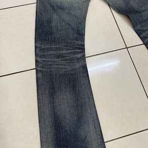 May include: Faded blue denim jeans with a classic five-pocket design. The jeans have a slight bootcut leg and show signs of wear, with subtle fading and whiskering details. The back pockets feature a signature stitch.