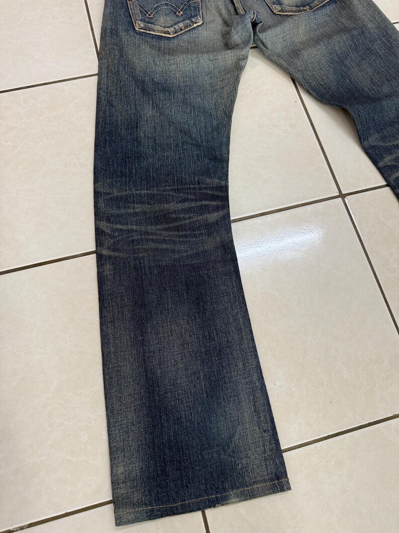 May include: Faded blue denim jeans with a classic five-pocket design. The jeans have a slight bootcut leg and show signs of wear, with subtle fading and whiskering details. The back pockets feature a signature stitch.
